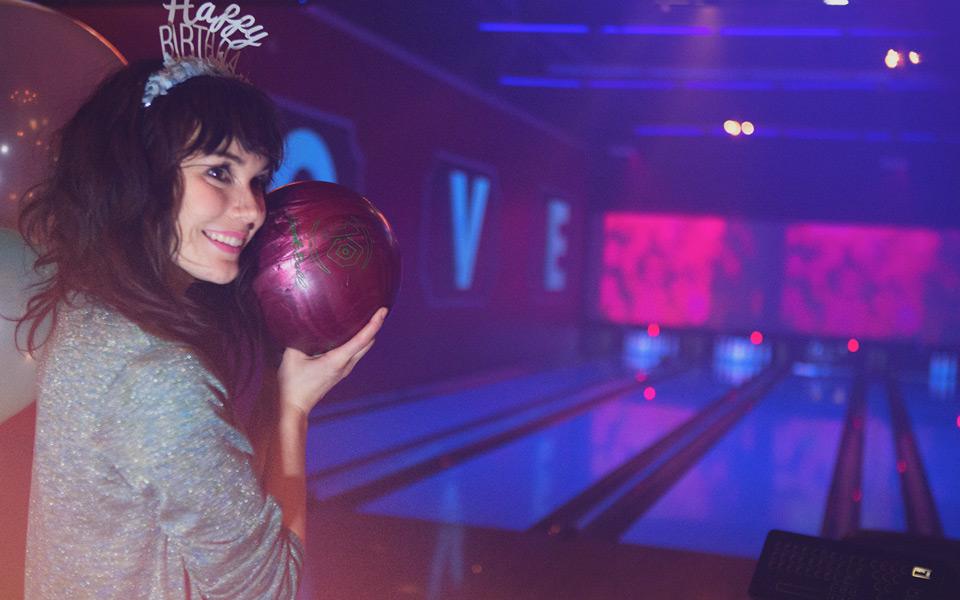 A woman smiling and holding a bowling ball in front of bowling lanes.