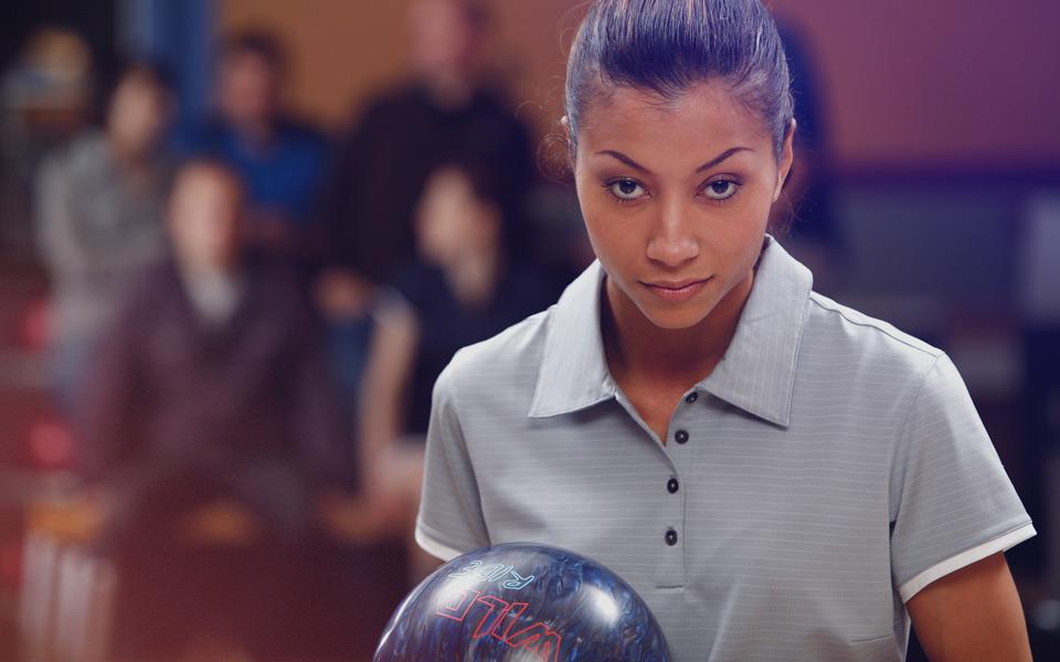 A woman holding a bowling ball in a bowling shirt staring straight at the camera. 