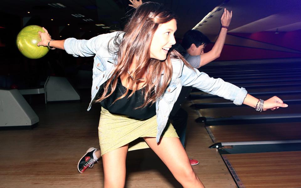 A girl bowling with a friend in the background. 