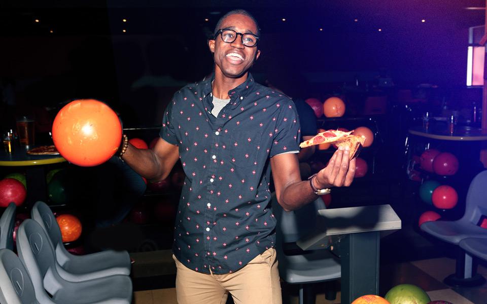 A man eating a slice of pizza while holding a bowling ball and laughing during a corporate party. 